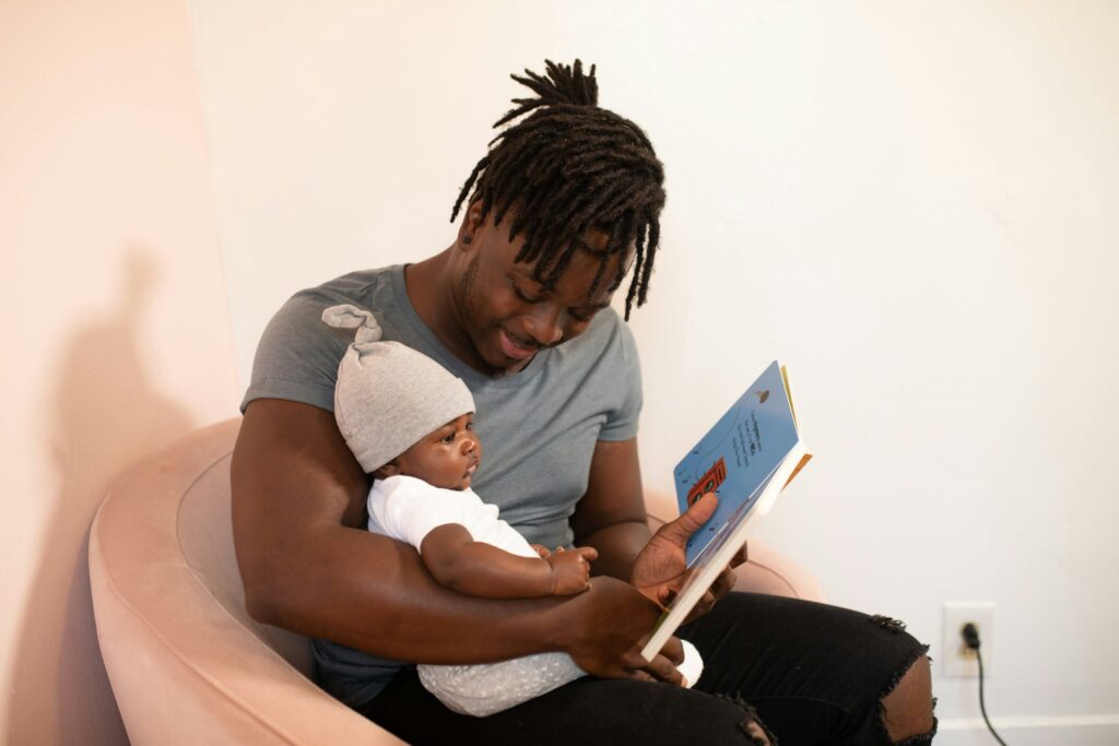 A young, Black father is reading a book to his infant child in their home