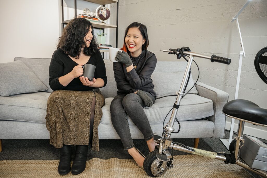 A Caucasian woman and her female Asian friend are enjoying coffee together on a grey couch at home. The Asian woman has her mobility scooter close by. Photo provided by Disabled and Here.