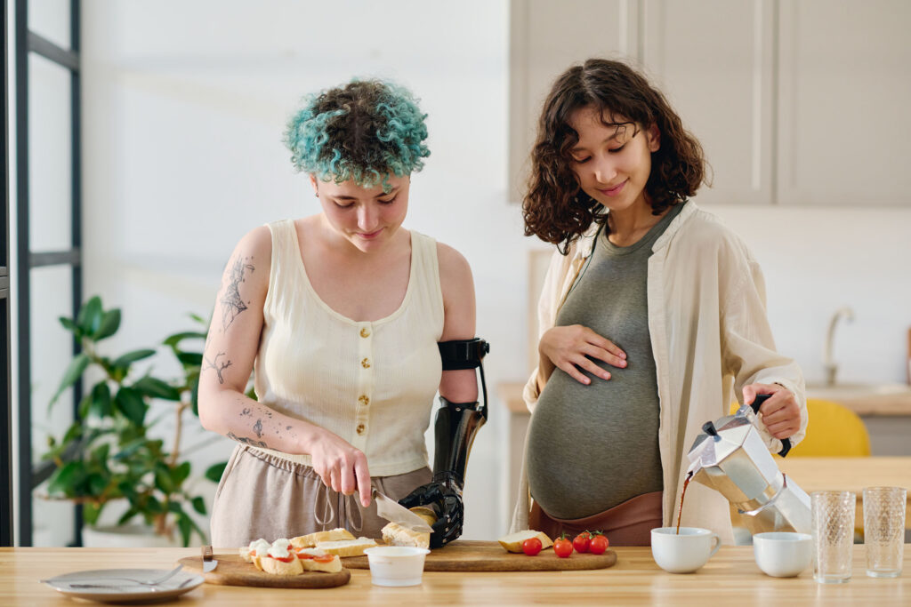 Two women are making breakfast together in their home. One woman is visibly pregnant and the other has a prosthetic arm