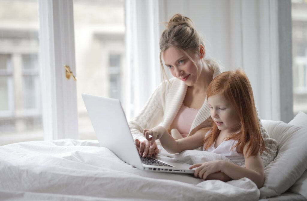 A mother and her daughter are relaxing in bed together. The mother is working on her advance healthcare directives on her laptop