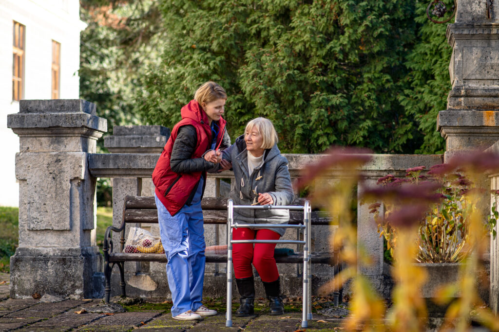 Senior woman with walker and her caregiver standing up from a bench in the park.