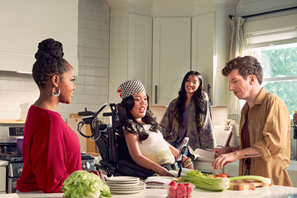 Four adults gather in a bright kitchen around a countertop with fresh vegetables and dishes. One person uses a wheelchair while participating in meal preparation with the group, as others stand nearby cutting produce and talking.